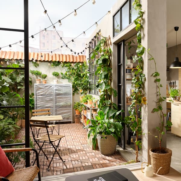 Terrasse avec caillebotis bois, plantes vertes à profusion, guirlandes lumineuses, table d’extérieur et ses deux chaises.