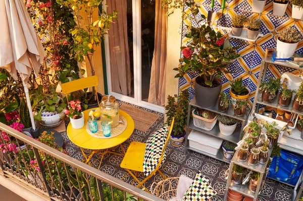 Terraza con conjunto de sillas y mesas amarillas de metal, sombrilla y muchas plantas distribuidas en el suelo, estanterías y colgadas. 