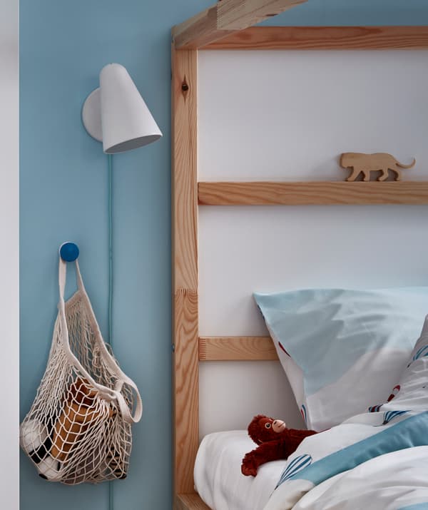 Tête de lit d’un lit d’enfant avec un filet rempli de peluches accroché à un bouton sur le côté. Une applique murale, une peluche à côté de l’oreiller.