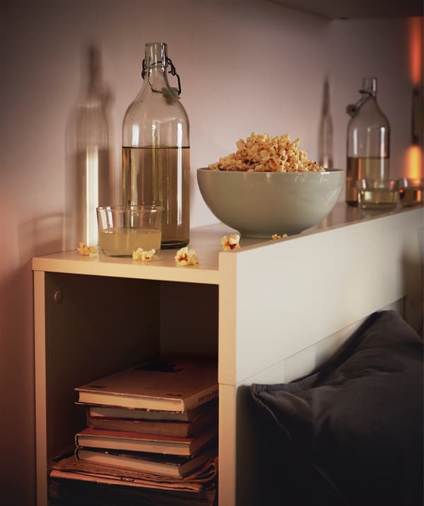 Tête de lit qui sert d’étagère, une pile de livres à l’intérieur, des bouteilles en verre remplies et un bol rempli de petites choses à grignoter posé au-dessus.