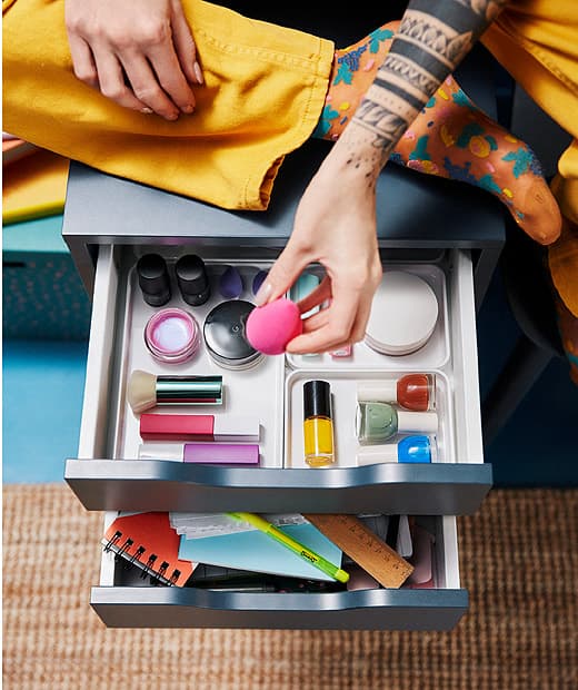 Tetovált lány ül egy fiókos szekrényen, sminkfelszerelésekkel.