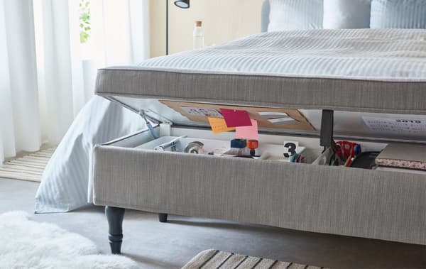 Textile-covered, combined bench and storage unit ajar at the foot end of a bed holding the contents of a typical home office.