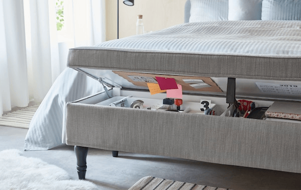 Textile-covered, combined bench and storage unit ajar at the foot end of a bed holding the contents of a typical home office.