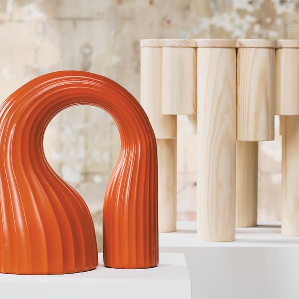 The arched and wavy decoration in eye-catching orange stands next to a wooden stool from the KONSTRUNDA collection.				