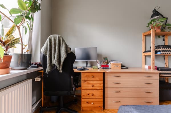 The bedroom before the makeover: a workspace by the window with a computer monitor, a black office chair and two wooden chests of drawers.