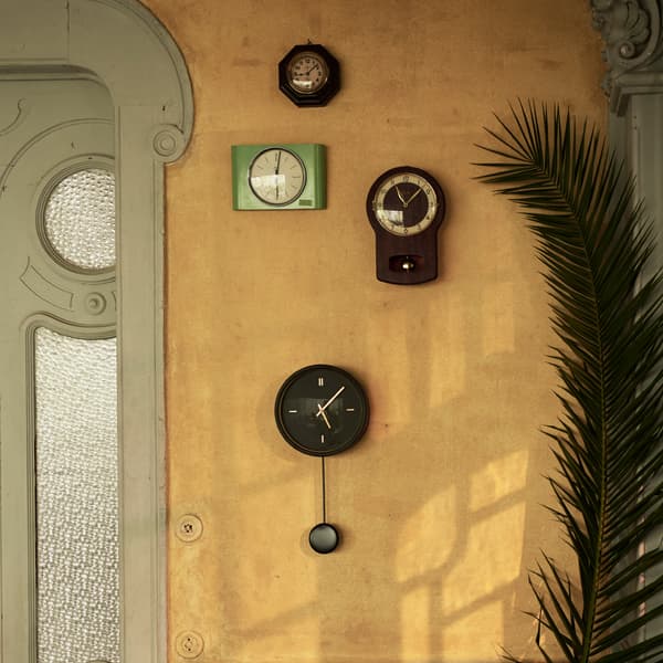 The black STURSK wall clock with gold details and a pendulum hanging from it. It's placed on a wall with old wall clocks.