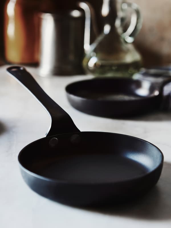 The black VARDAGEN frying pan placed on a grey marble countertop with a frying pan and kitchen accessories at the back.