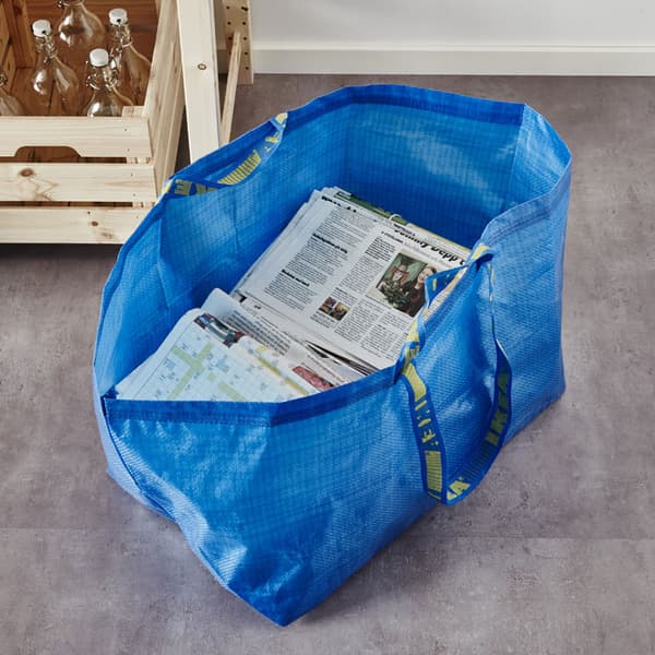 The blue FRAKTA bag sitting on the ground with newspapers inside.
