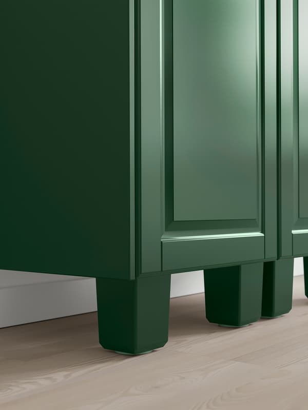 The bottom corner of a dark-green kitchen cabinet with matching dark-green legs. The door has a frame and a bevelled panel.