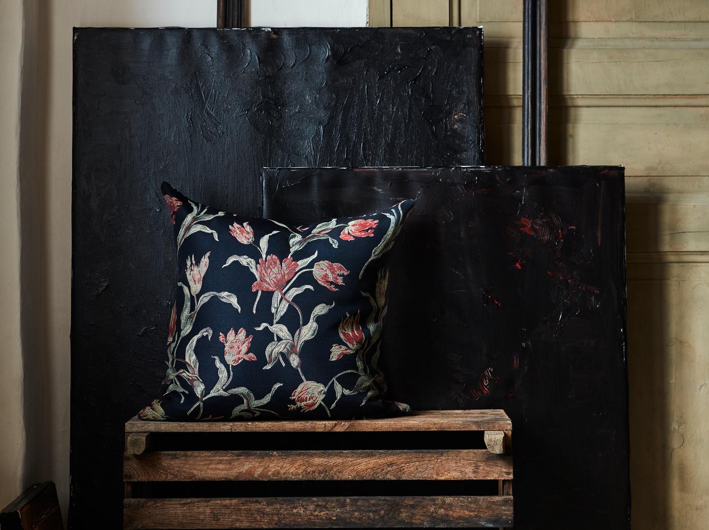 The hand-embroidered ÅLANDSROT cushion with a tulip pattern on a dark-blue background placed on wooden crates in a dark room.