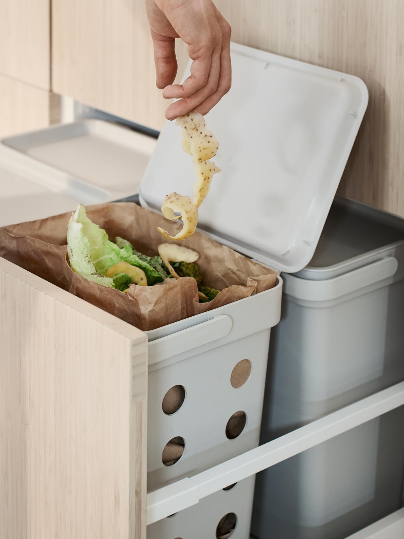 The hand of a person lowering a long string of potato peel into a HÅLLBAR bin containing organic kitchen waste.