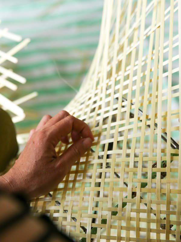 The hand of an artisan that is weaving a light, yellowish bamboo lattice by hand onto a metallic lamp base.