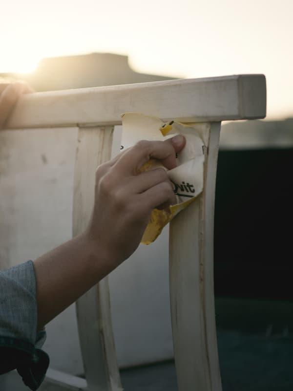The hand of someone with rolled up sleeves sanding the back of a white wooden chair whilst outside in the evening sun.
