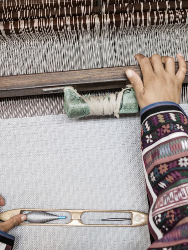 The hands of a weaver wearing a traditionally patterned top in the process of weaving a white rug on a loom.