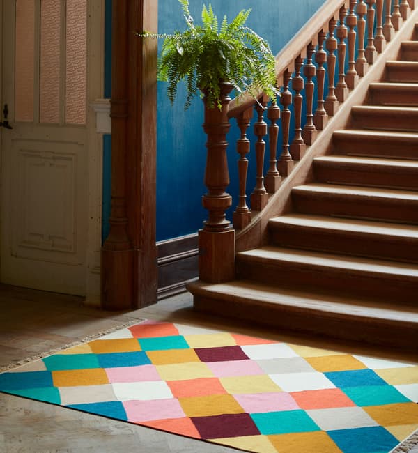 The handwoven VINDERÖD rug in wool made in a colourful, chequered pattern, placed at the bottom of a staircase.