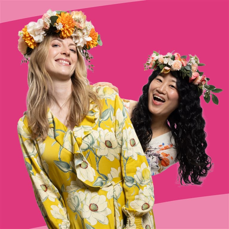 The image features two smiling women wearing floral crowns against a vibrant pink background. The woman in the foreground has blonde hair and is wearing a yellow dress with white floral patterns, while the woman behind her has dark, curly hair and is wearing a lighter-colored floral top. Both appear joyful.