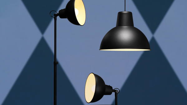 The image highlights the IKEA SKURUP lamp series, showcasing its industrial and functional design. It features three lamps: a floor lamp, a table lamp, and a pendant lamp, all in matte black with dome-shaped shades. The strong, direct lighting creates distinct, geometric shadows on the blue background, emphasizing the lamps' practical and stylish aesthetic. The SKURUP series is ideal for modern, minimalist interiors, providing focused illumination and a sleek look.