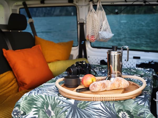 The inside of the van with a view of the dining table with a jungle print tablecloth and a loaf of bread, an apple and a coffee maker layed out on a wooden tray 