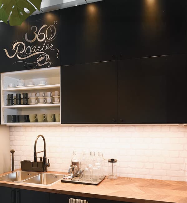 The kitchen area of the café showcasing a sink on a wooden top as well as black cabinet with cups. 