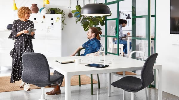The man sitting on YPPERLIG chair with armrest is talking with woman holding notebook in the office where has BAKANT table and LÅNGFJÄLL office swivel chair