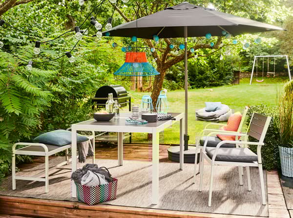 The modern, outdoor SJÄLLAND table, chairs and bench in dark grey on a terrace with a parasol, overlooking a lawn.