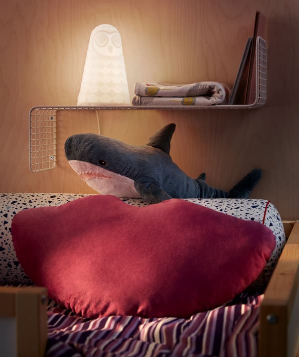 The pillow end of at loft bed with cushion, cuddly toy shark and a slimline wall shelf holding a night light, books, quilt.