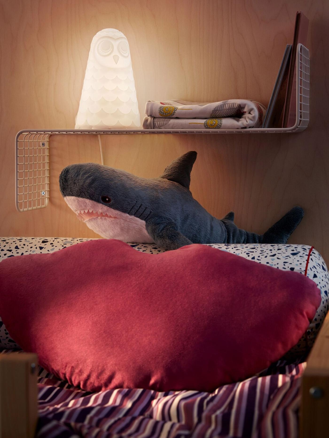 The pillow end of at loft bed with cushion, cuddly toy shark and a slimline wall shelf holding a night light, books, quilt.