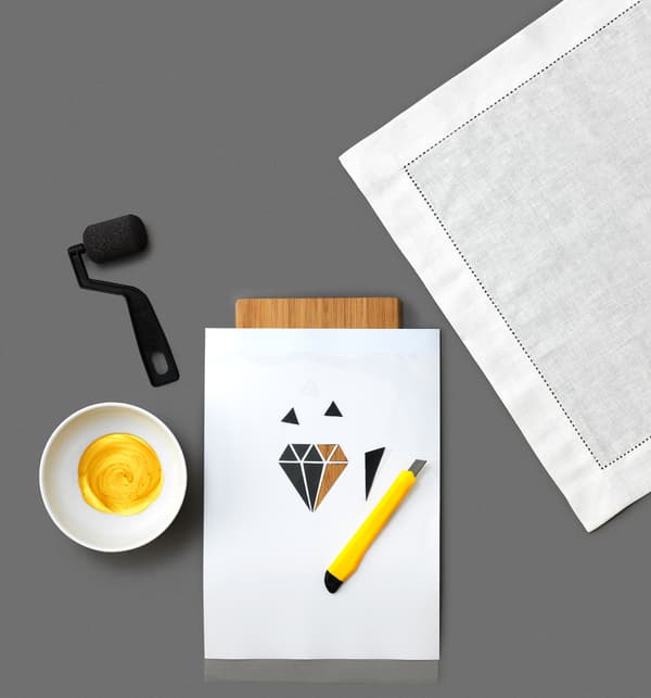 The second step of painting fabric stencils on a GULLMAJ white cloth napkin from IKEA is to cut the stencil. Put the patterned paper and plastic sheet on a wooden chopping board. Cut with a utility knife.