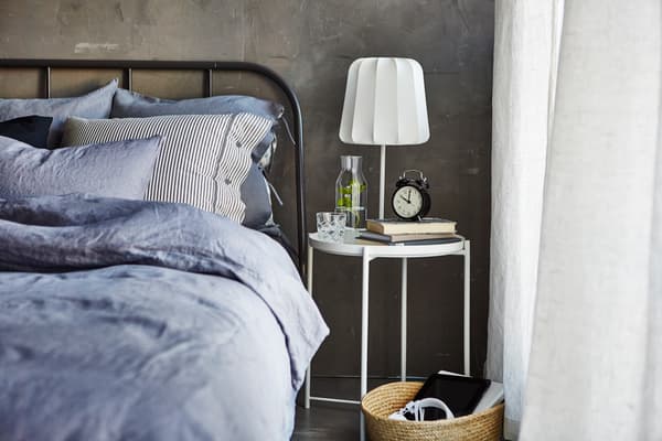 The side of a made bed with a bedside table, glass of water, books, an alarm clock and dimmable lamp.