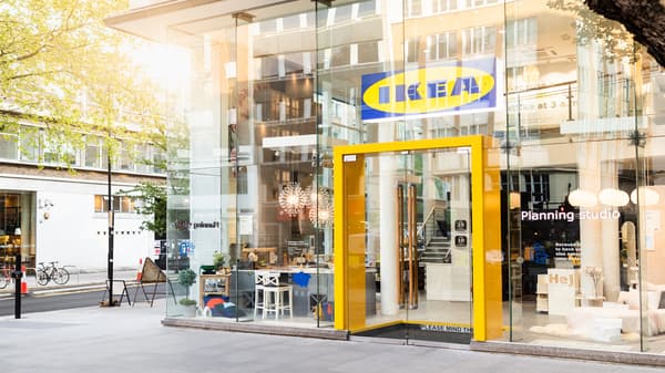 The sun is shining through the glass shopfront of an inner-city IKEA store. The entrance is framed in bright yellow.