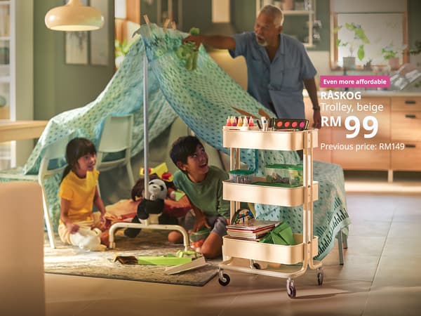 The visual shows a beige RÅSKOG trolley filled with art and craft supplies in a living room, while two children play inside a blanket fort and an older man helps build it in the background. The text reads: “Even more affordable. RÅSKOG trolley, beige, RM99. Previous price: RM149.”