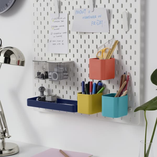 The white SKADIS pegboard on a wall with desk accessories and papers displayed on it.