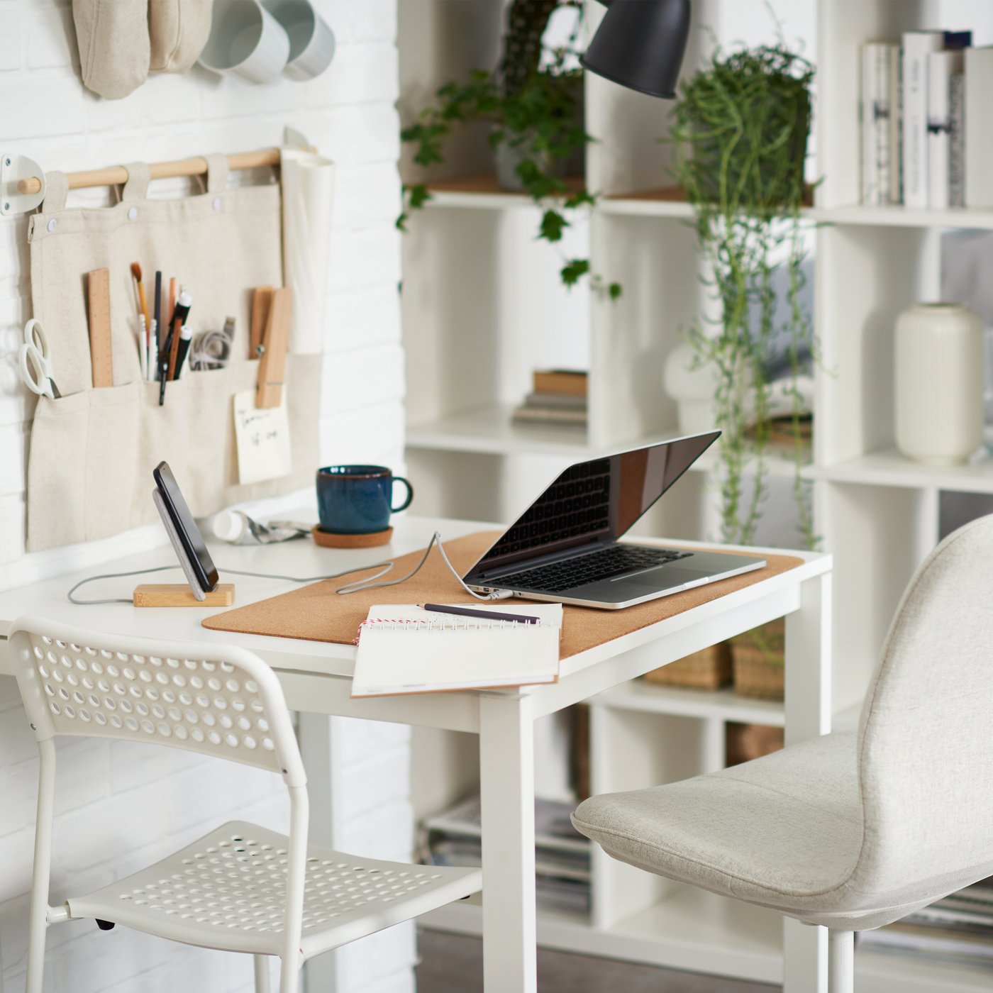 There is a laptop on a white table, and there is one white and one beige chair at the table. Next to it is a shelf with books, plants, and storage items.