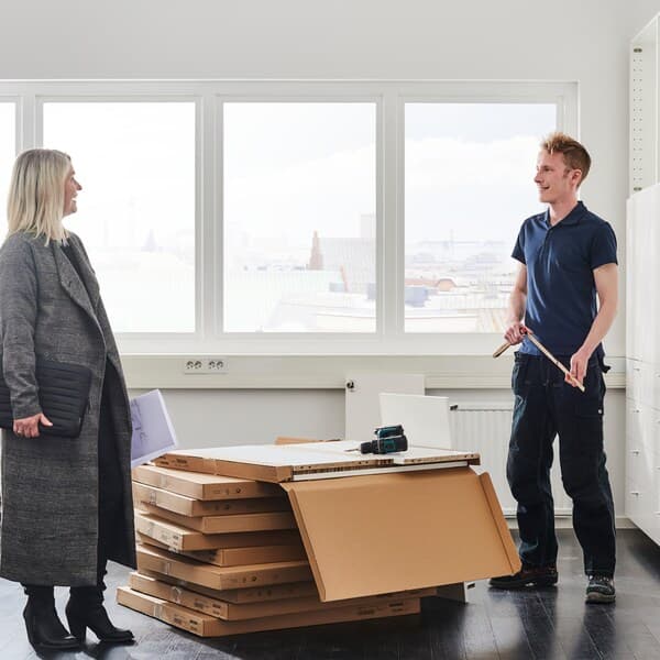 Thin boxes of flatpacked furniture stacked in between a man with a measuring stick and a woman in a grey coat.