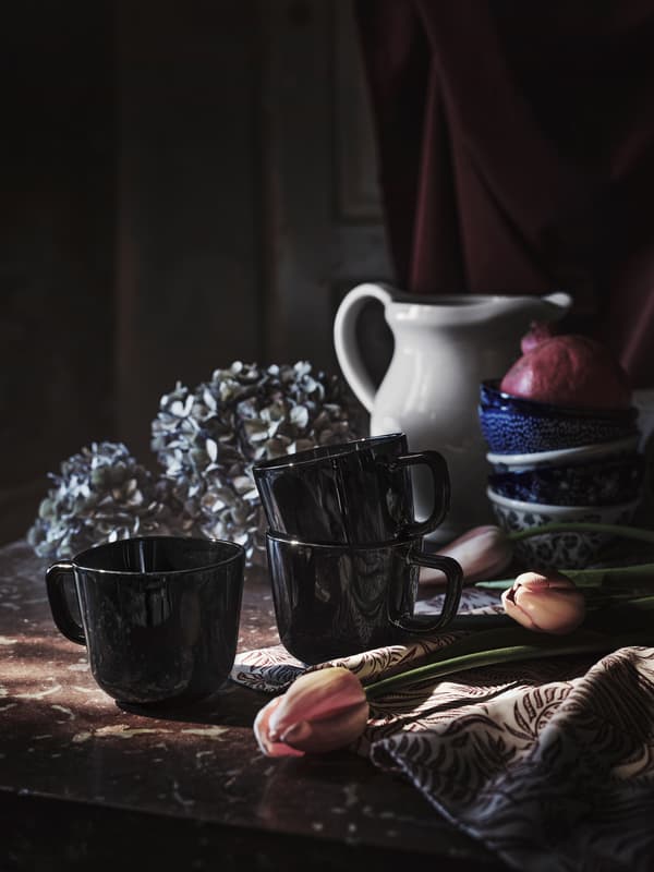 Three BACKIG mugs in glossy black glass placed on a table with other mugs, a jug and some tulips lying on the surface.