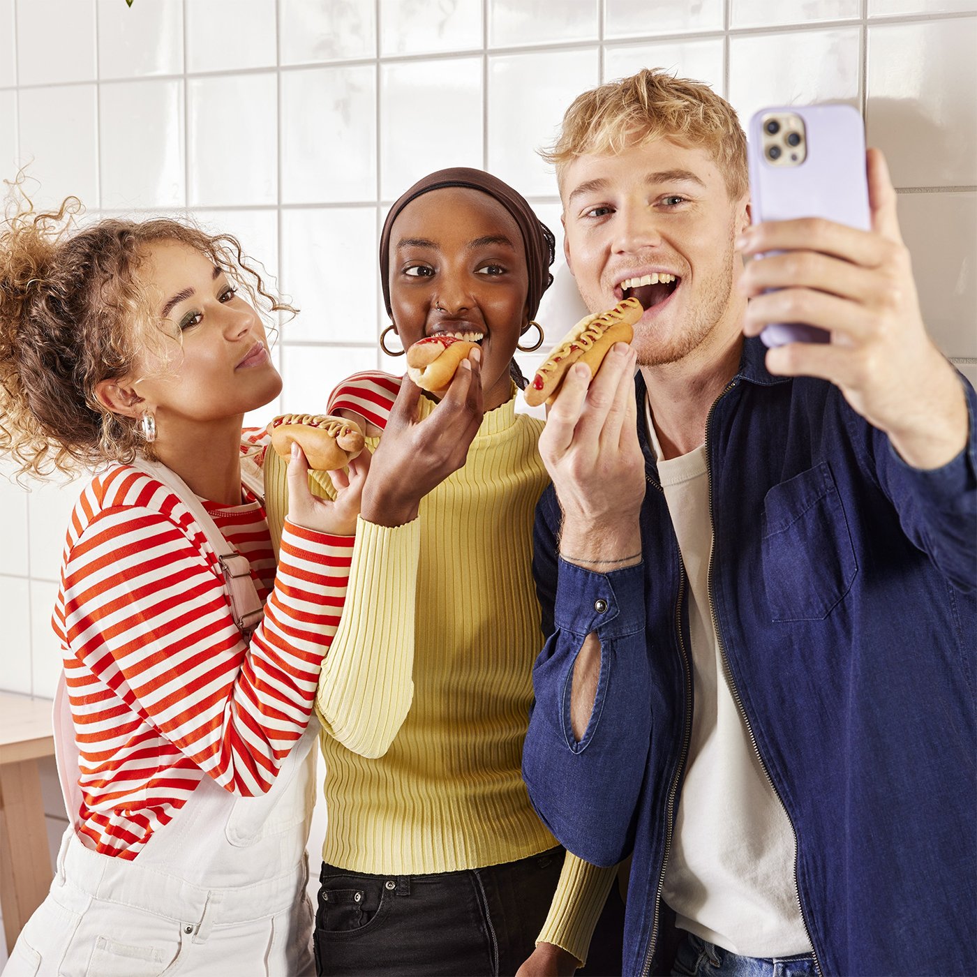 Three cheerful teenagers in colourful clothes taking a selfie while holding hot dogs.