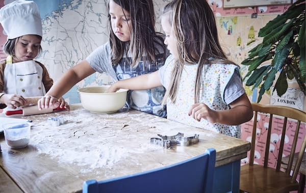 Three children standing around a table with flour, dough and baking utensils.