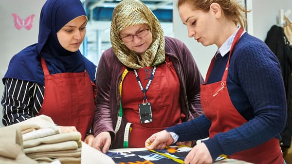 Three co-workers from Yalla Trappan sewing studio in Malmö, Sweden, measuring some fabric while wearing red aprons.