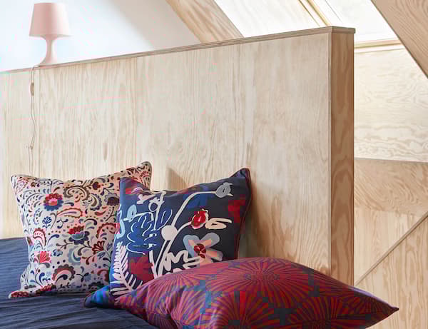 Three colorful pillows with flower patterns against a wood wall with a pink lamp.
