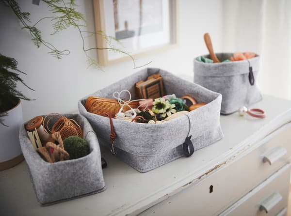 Three decorative, grey felt containers in different sizes filled with knick-knacks, standing on a vintage cabinet.