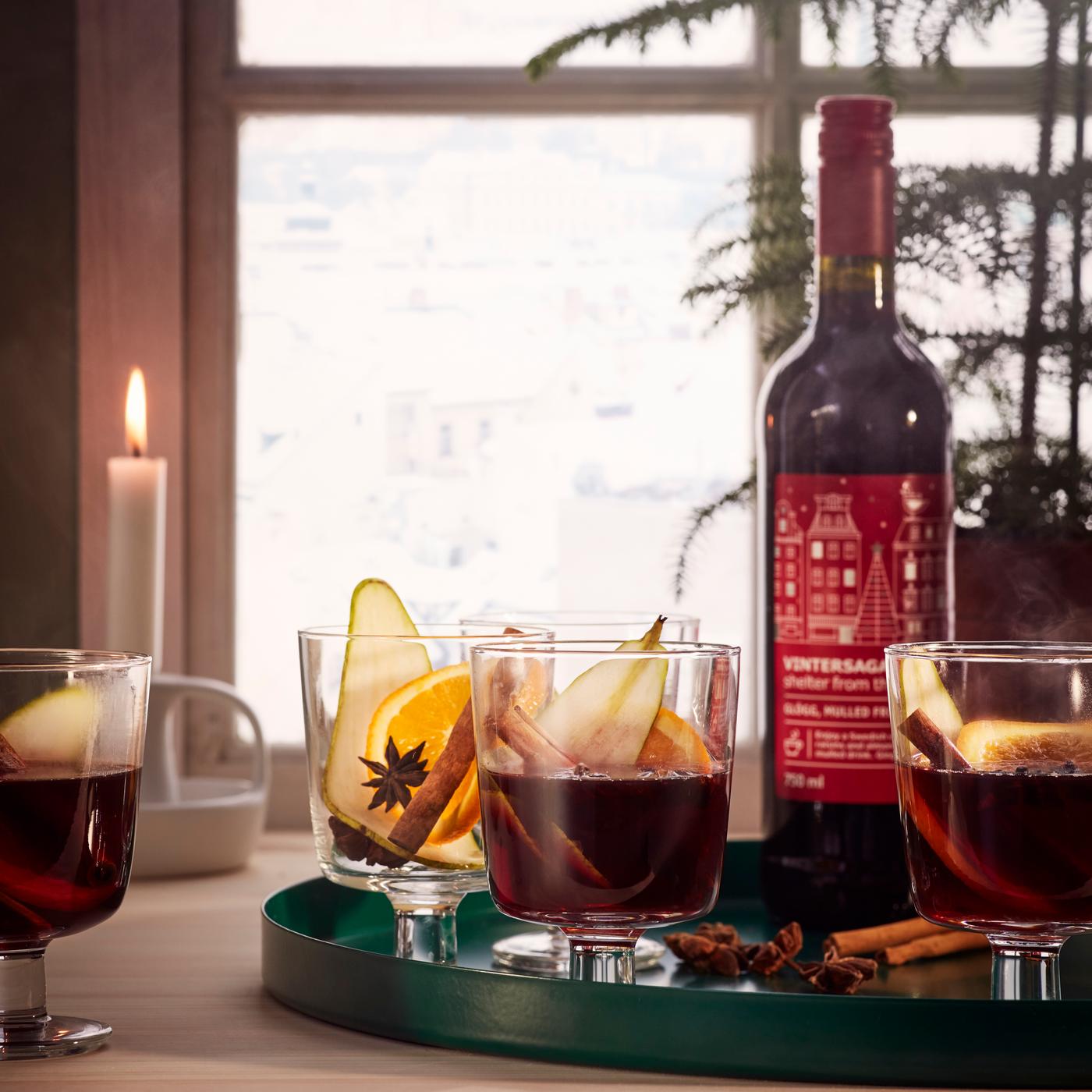 Three glasses and a bottle of VINTERSAGA glögg, mulled fruit drink, placed on a green serving tray with a candle lit in the background.