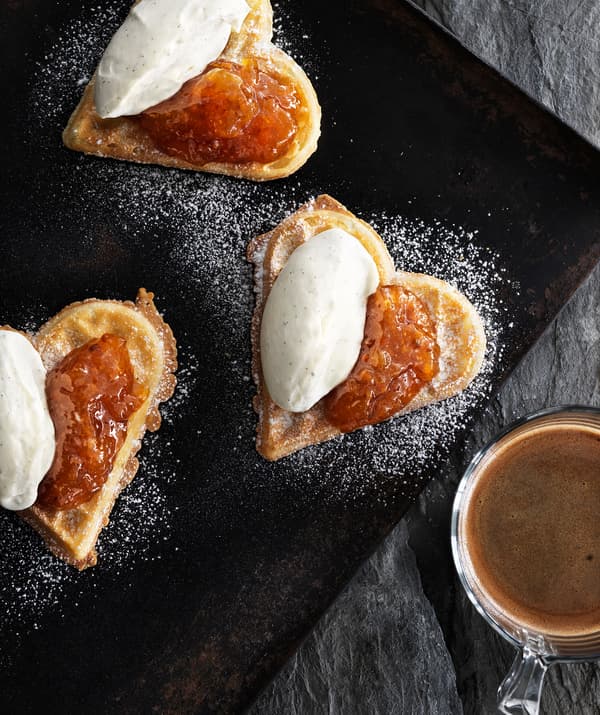 Three heart-shaped waffles with jam and whipped cream on a baking tray, with coffee in a glass to one side.