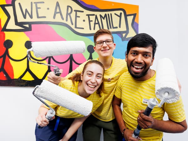 Three IKEA co-workers in yellow tshirts with paint rollers. We are family sign in the background