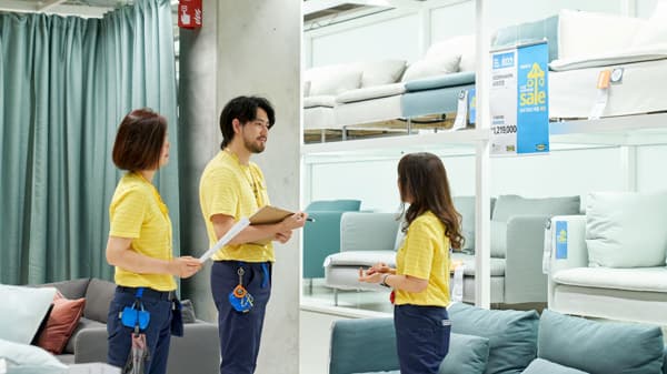 Three IKEA co-workers in yellow uniforms in the sofa area, reviewing layout and signage with a clipboard and notes.