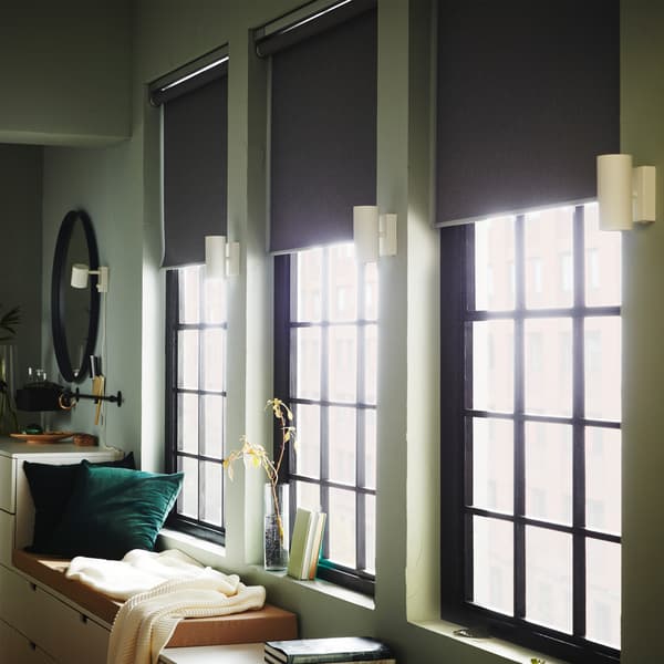 Three large windows that are slightly covered by grey block-out roller blinds, four white wall lamps and two green cushions.
