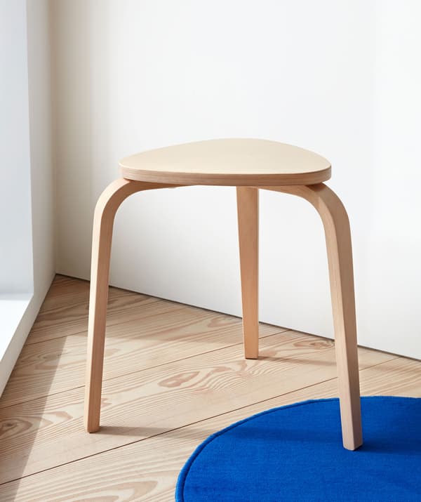 Three-legged, pale wood stool standing in a corner on a wooden floor, with one leg on a small blue rug.