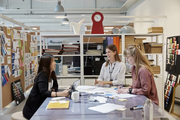 Three people are having a discussion in an office at a table with many pieces of paper scattered around.