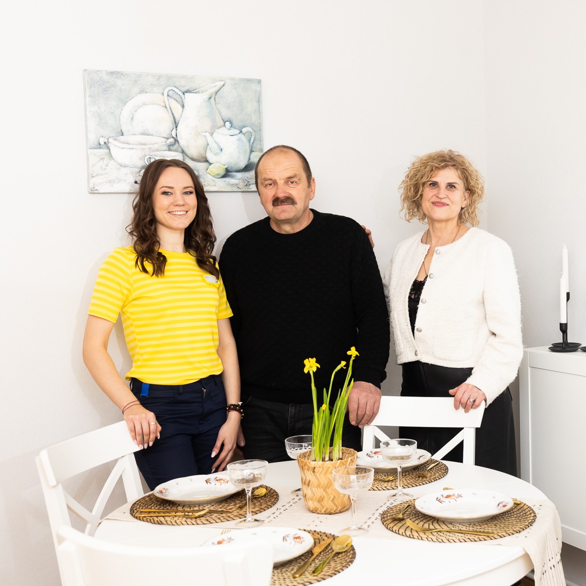 Three people stand in a bright, minimalist dining area next to a round table set with plates. A white sideboard and a round wall mirror are visible, creating a cosy, tidy interior atmosphere.