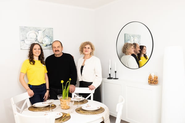 Three people stand in a bright, minimalist dining area next to a round table set with plates. A white sideboard and a round wall mirror are visible, creating a cosy, tidy interior atmosphere.