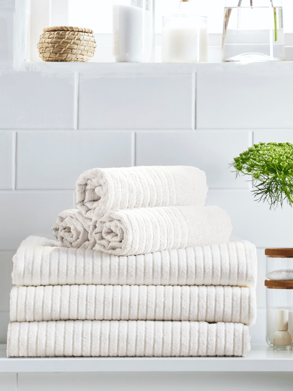 Three rolled-up towels are on top of a pile of three folded towels on a white surface in a tiled bathroom.